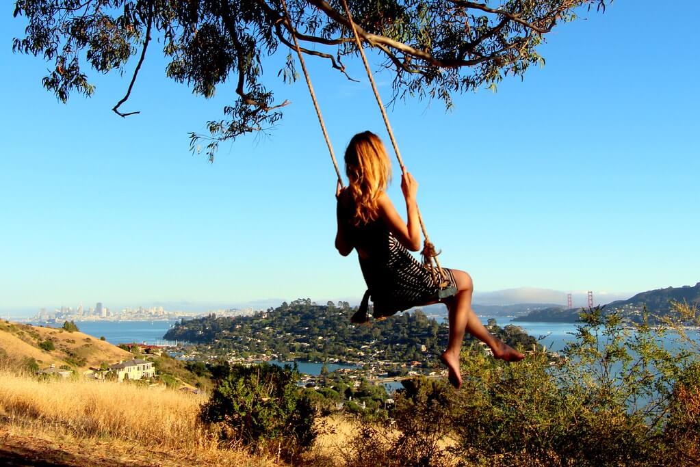 A woman in dress is swinging on the Hippie Tree Swing with nice view of San Francisco on a sunny day.