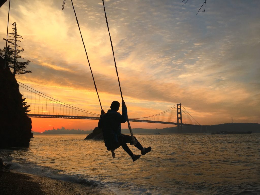 Silhouette of a person on the Kirby Cove Swing with a vibrant sunset behind the Golden Gate Bridge and the San Francisco skyline.