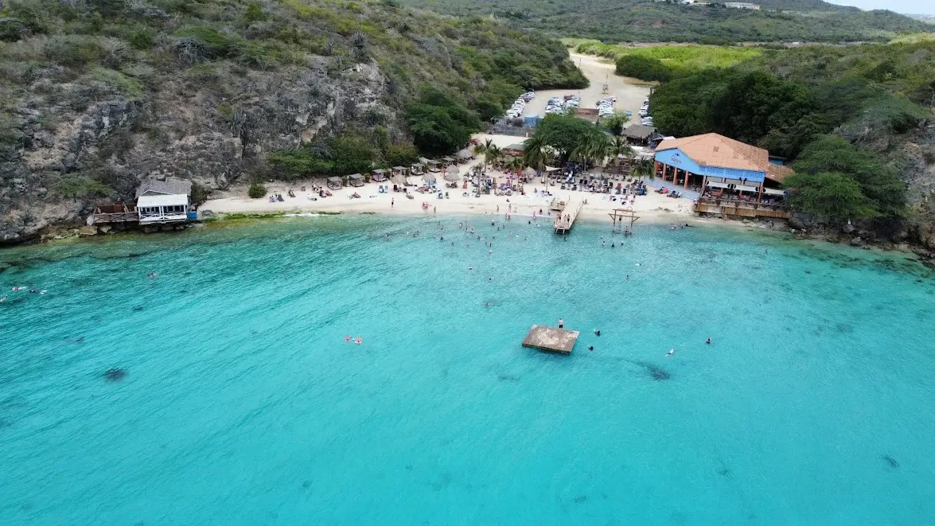 Aerial view of Kokomo Beach with a swing in the ocean, beachgoers relaxing on the shore, and a beachside restaurant surrounded by tropical scenery.