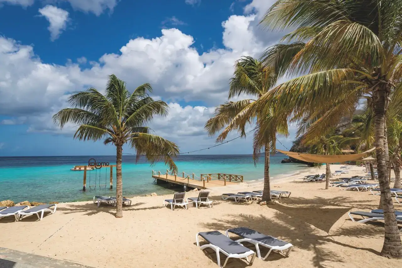 A tropical beach scene with palm trees, white sand, and turquoise water, featuring lounge chairs, a wooden pier, and a sign reading "Kokomo" with swings in the water.