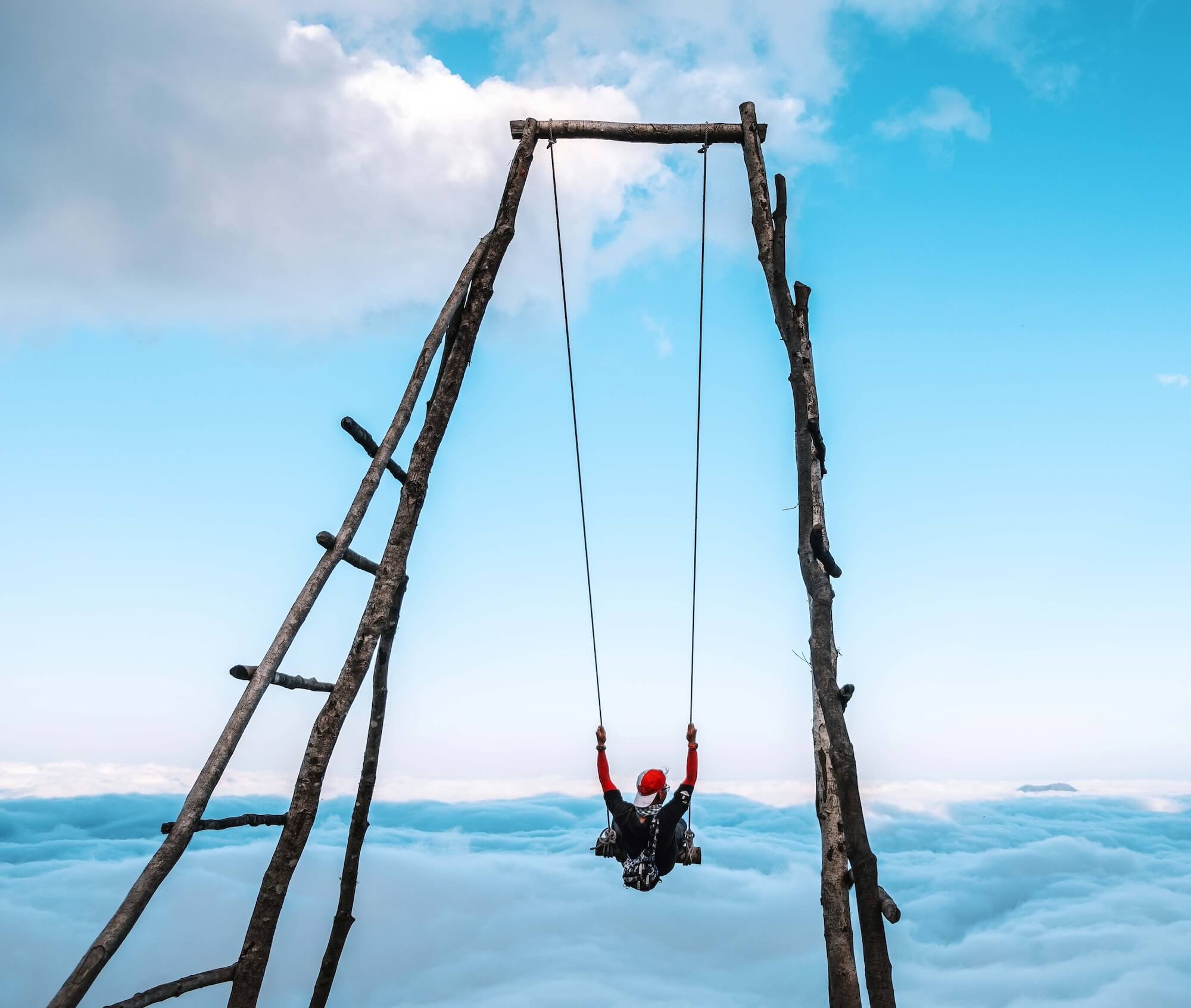 A person swings high above a sea of clouds on a tall wooden swing structure against a bright blue sky.