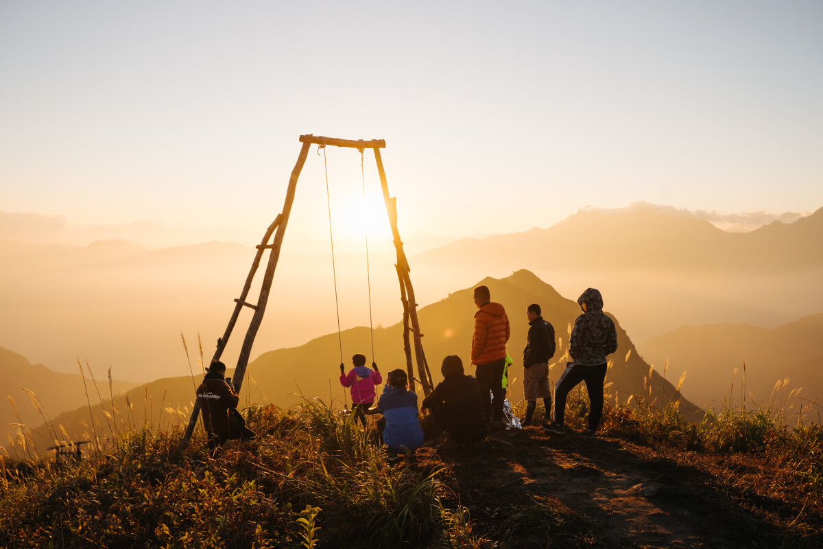 Silhouetted figures gather around a wooden swing frame at sunset, with one person swinging above misty mountain peaks.