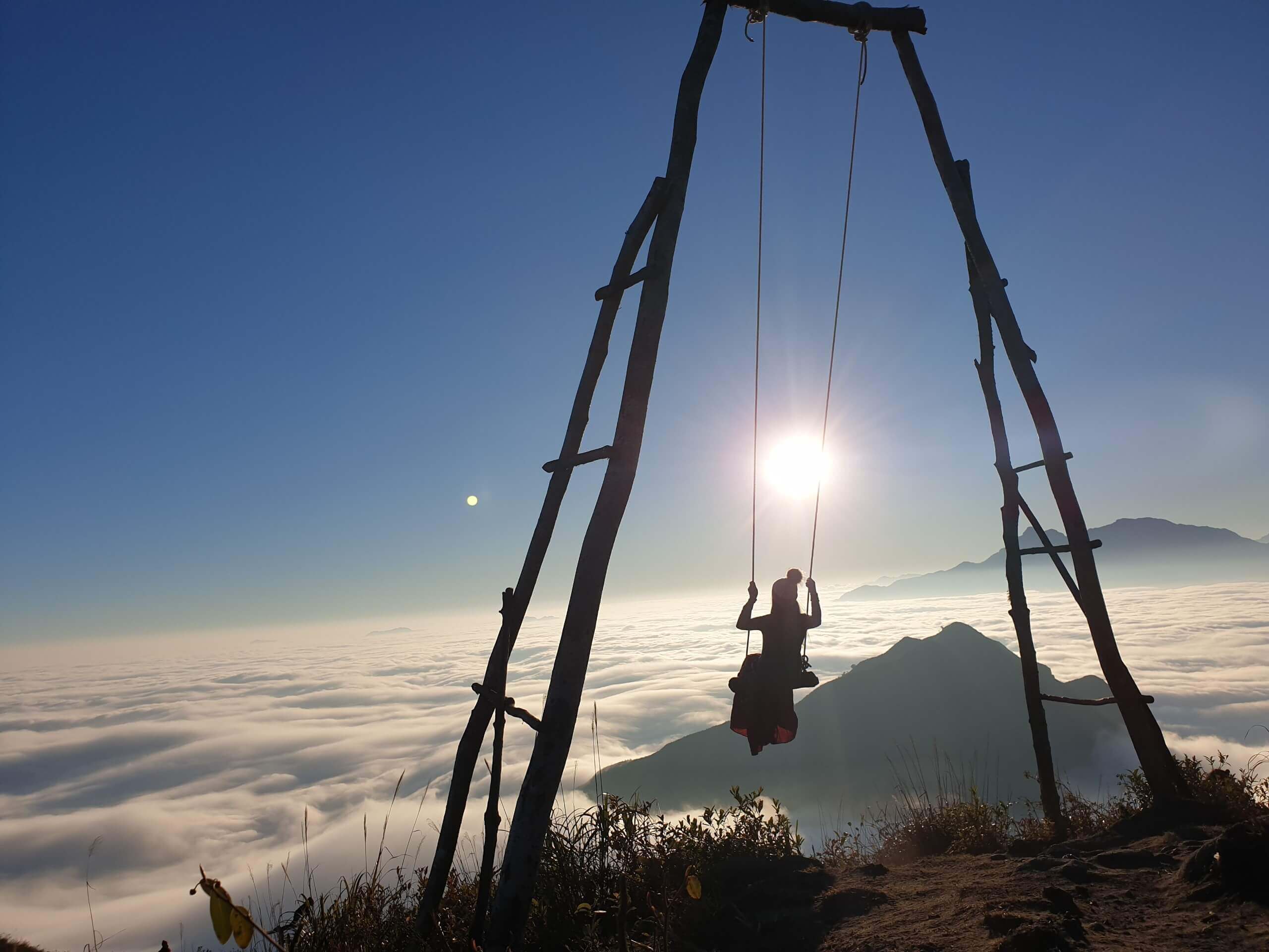 A silhouetted woman swings above a sea of clouds at sunrise, framed by a tall wooden swing structure and distant mountains.