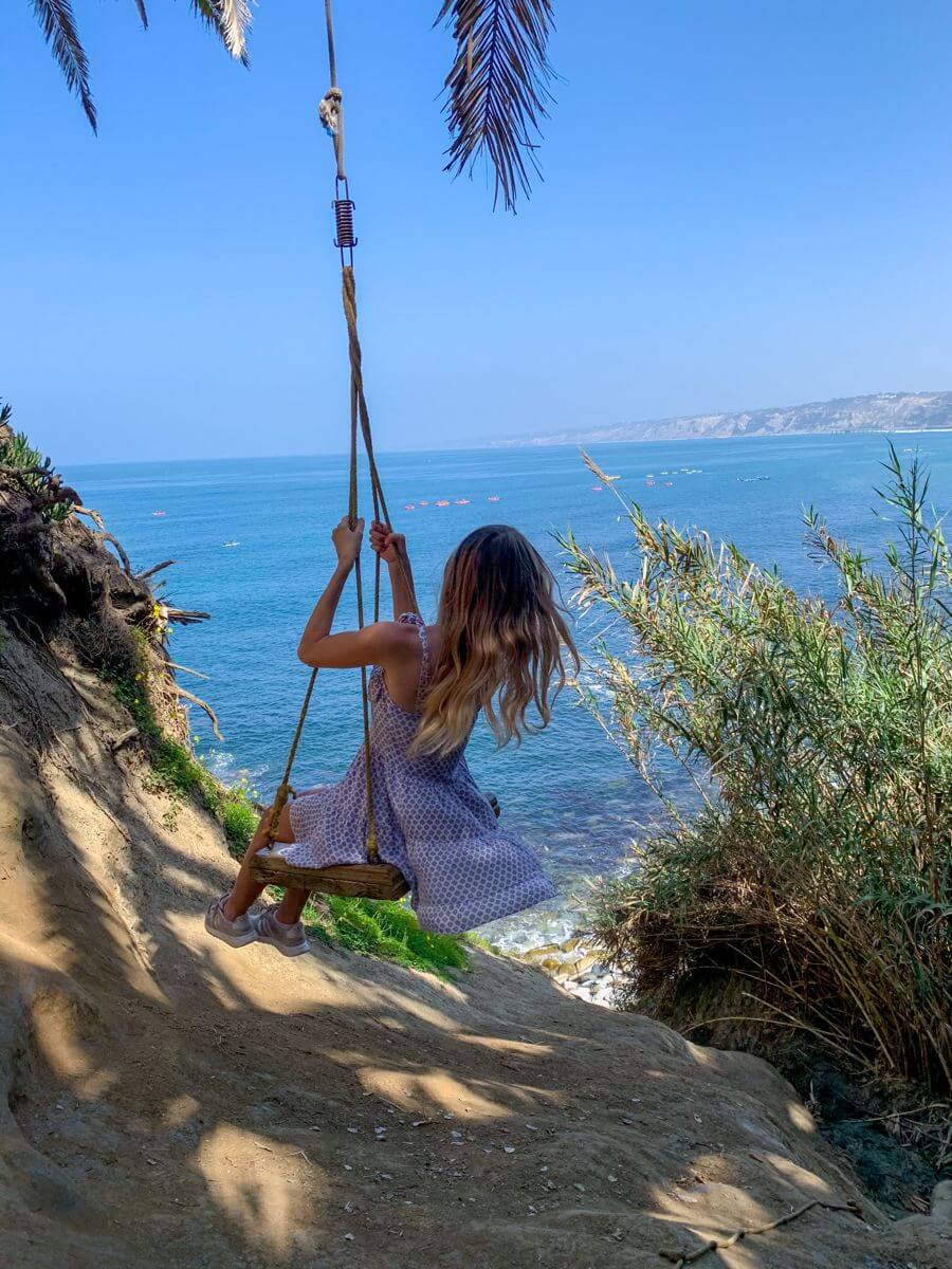 A woman swings on a La Jolla Cove Swing overlooking the ocean, with a coastal cliff, palm trees, a blue sea, and distant coastline visible in the background.