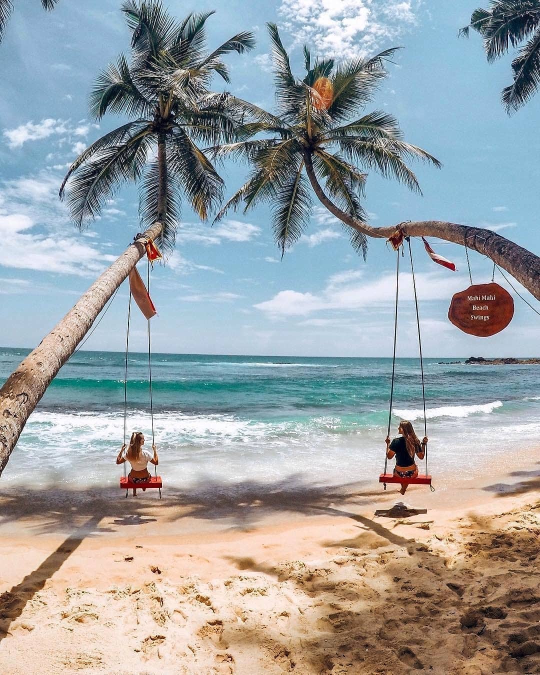 Two people enjoy red swings suspended from leaning palm trees on a picturesque tropical beach with turquoise waters, white sand, and a "Mahi Mahi Beach Swings" sign visible.