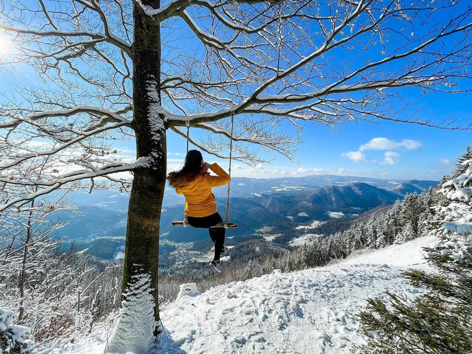 A person sits on a swing tied to a snow-covered tree, gazing at a breathtaking winter landscape of snow-capped mountains under a bright blue sky.