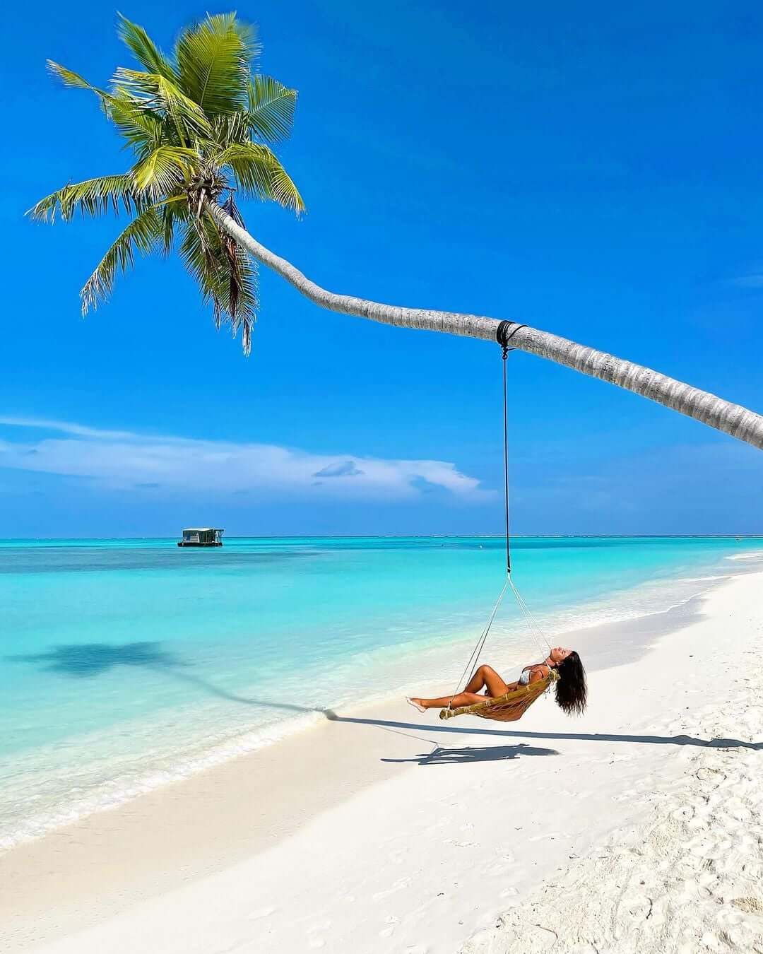 A woman lounges on a swing suspended from a palm tree on a white sandy beach, with crystal-clear turquoise waters and a bright blue sky in the background.