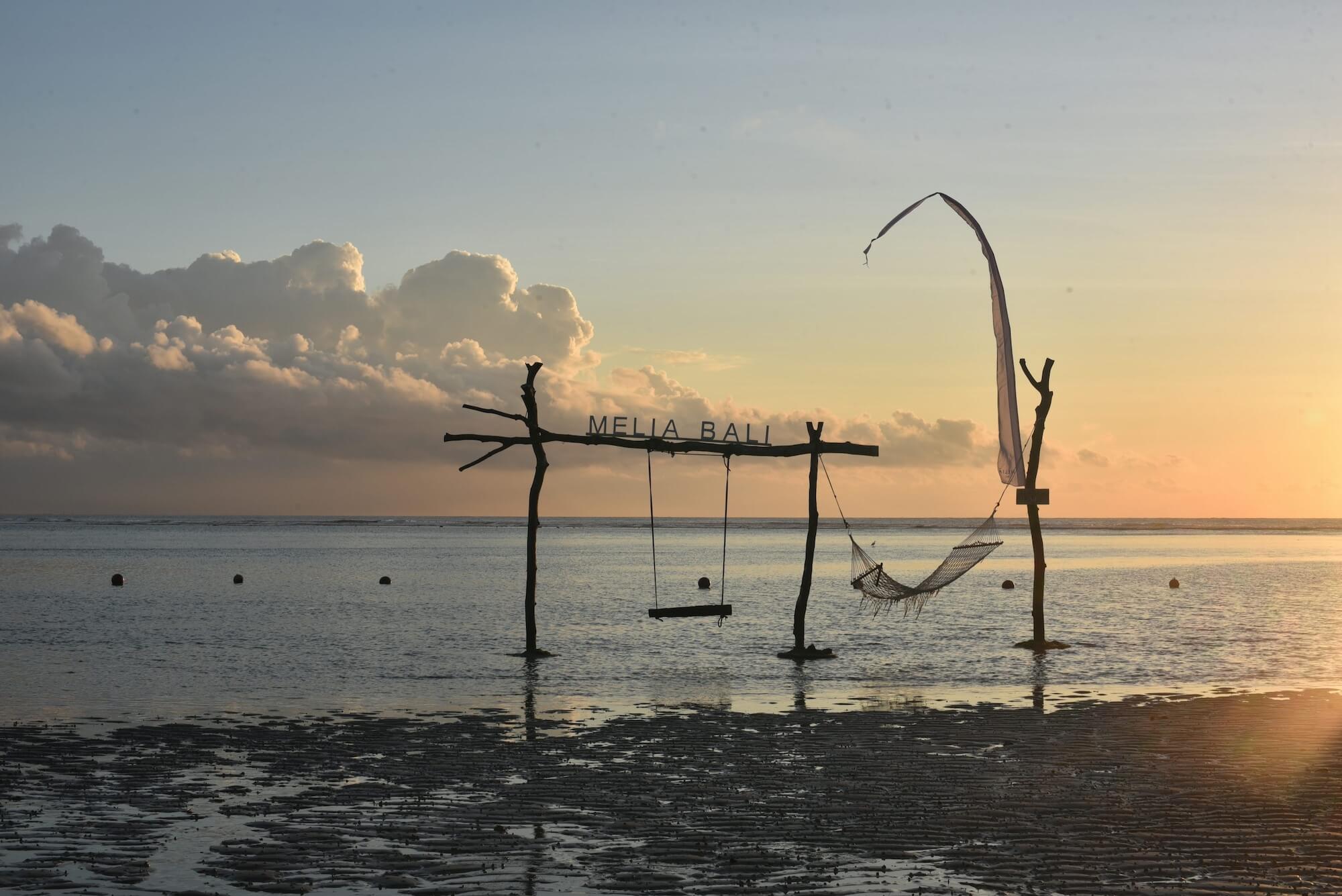 Wooden structure with "MELIA BALI" sign, swing, and hammock silhouetted against golden sky, calm sea, and fluffy clouds on horizon.