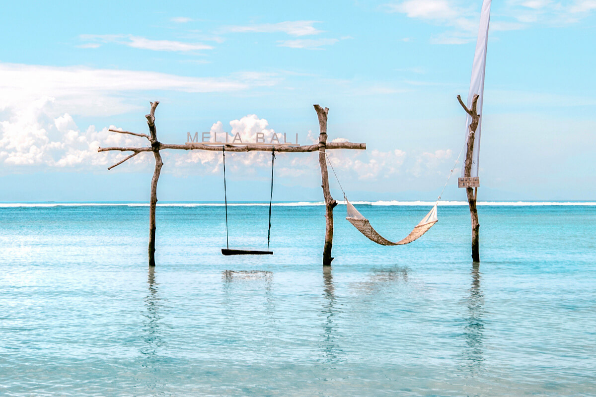 A rustic wooden frame with a swing and hammock suspended over crystal-clear turquoise waters, inviting relaxation with a view of the endless ocean horizon.