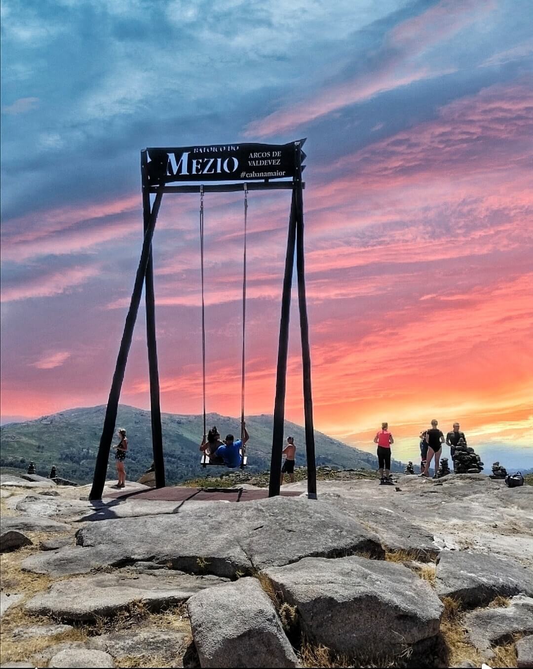 People gather around the Baloico do Mezio swing with a stunning sunset backdrop of pink and blue skies in Arcos de Valdevez, Portugal.