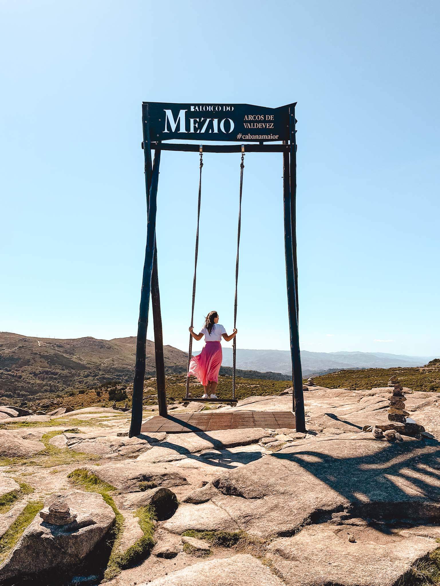 A woman in a flowing pink skirt enjoys the view from the Baloico do Mezio swing, surrounded by rocky terrain and rolling hills on a bright, clear day.