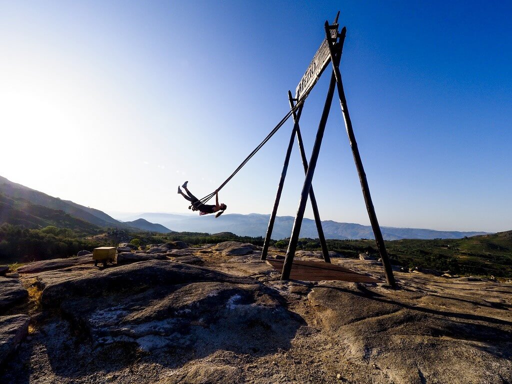 A person swings on a giant swing structure atop a rocky mountain peak, silhouetted against a clear blue sky with distant mountains in the background.