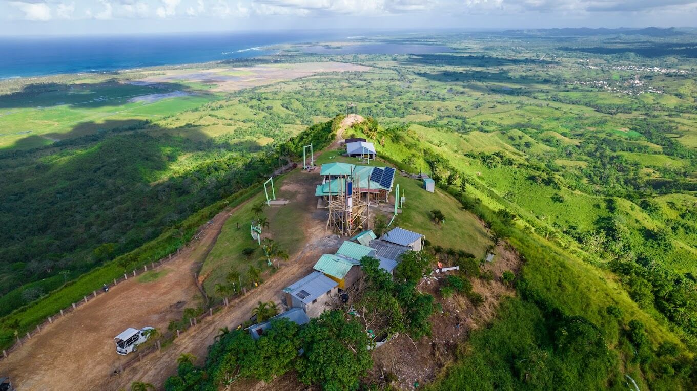 An aerial view of Montana Redonda hilltop small buildings and solar panels overlooking lush green landscape and distant coastline.