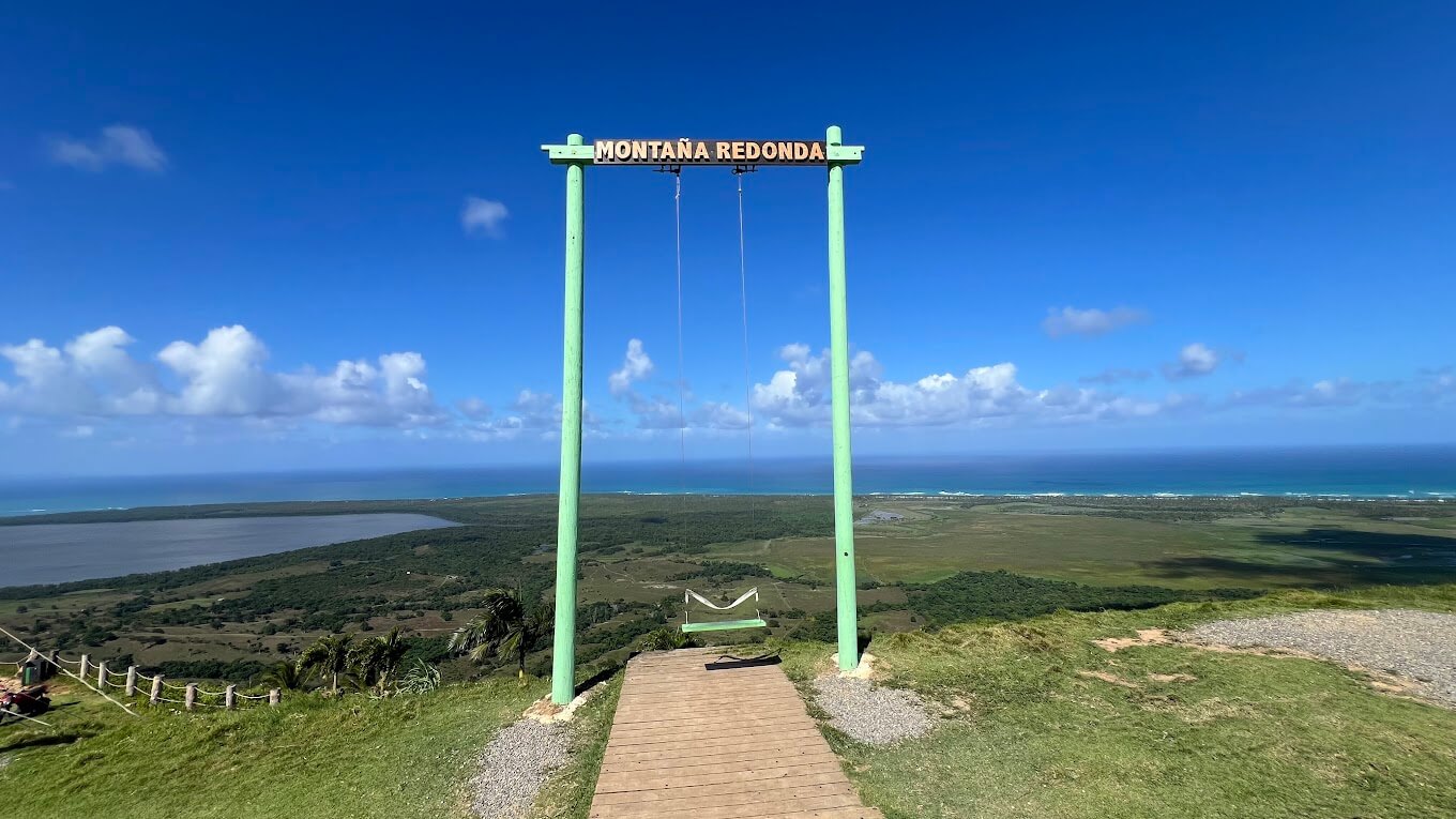 A large green swing frame reading "Montana Redonda" stands atop a hill, offering panoramic views of the coastline and landscape below.