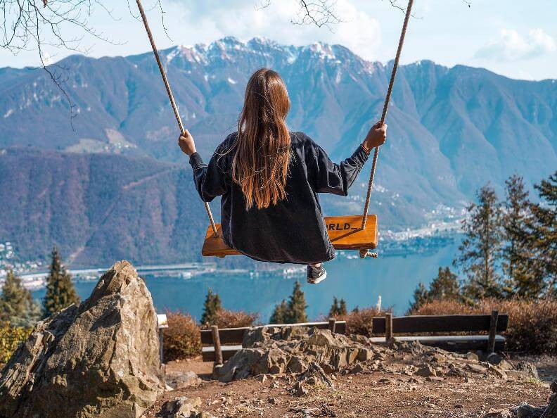 A girl with long brown hair swinging on a wooden swing overlooking a scenic mountain range and a lake at Parco San Grato.