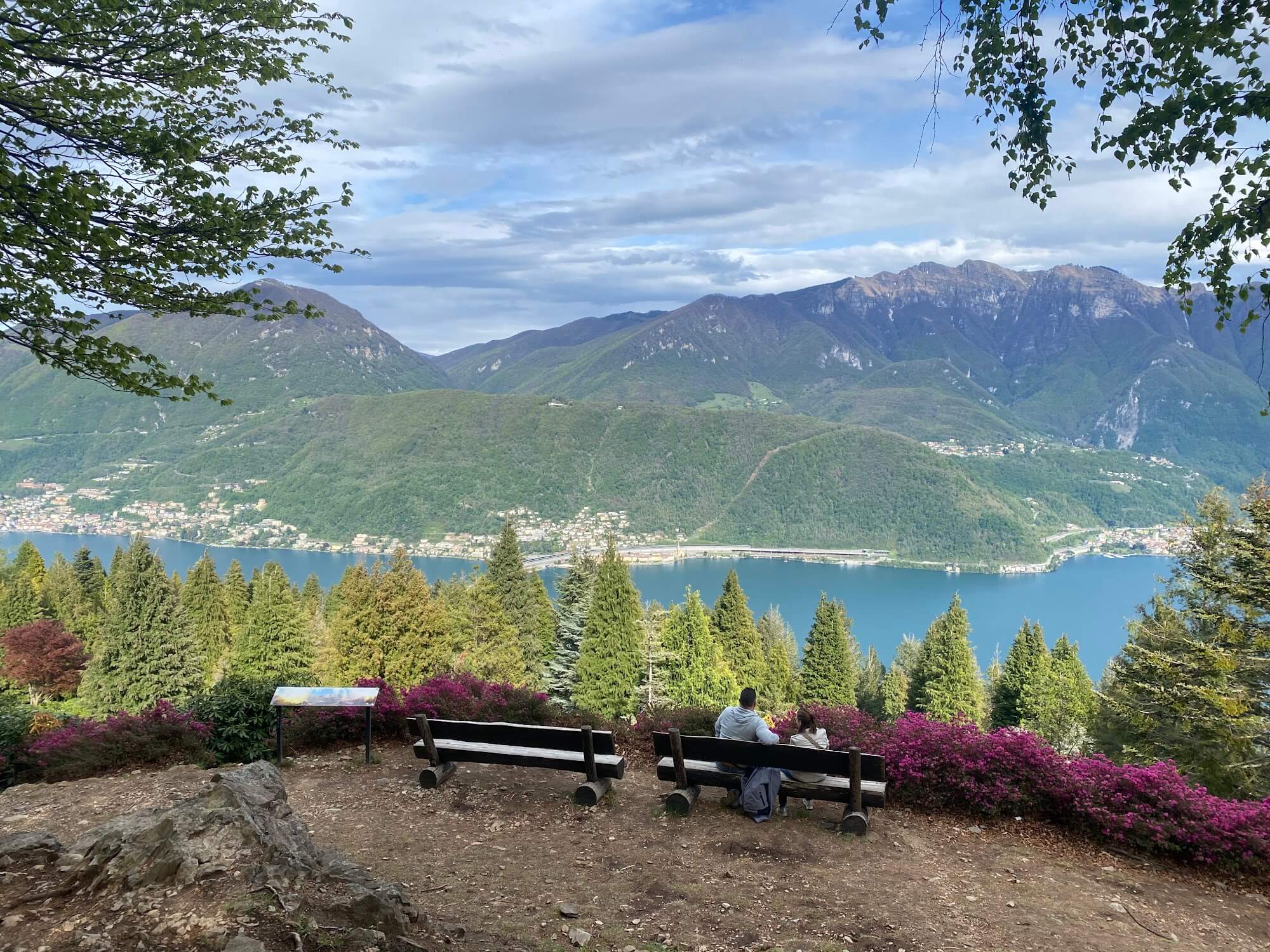 A panoramic view from a hilltop at Parco San Grato with benches overlooking a lush green valley, a lake, and mountains.