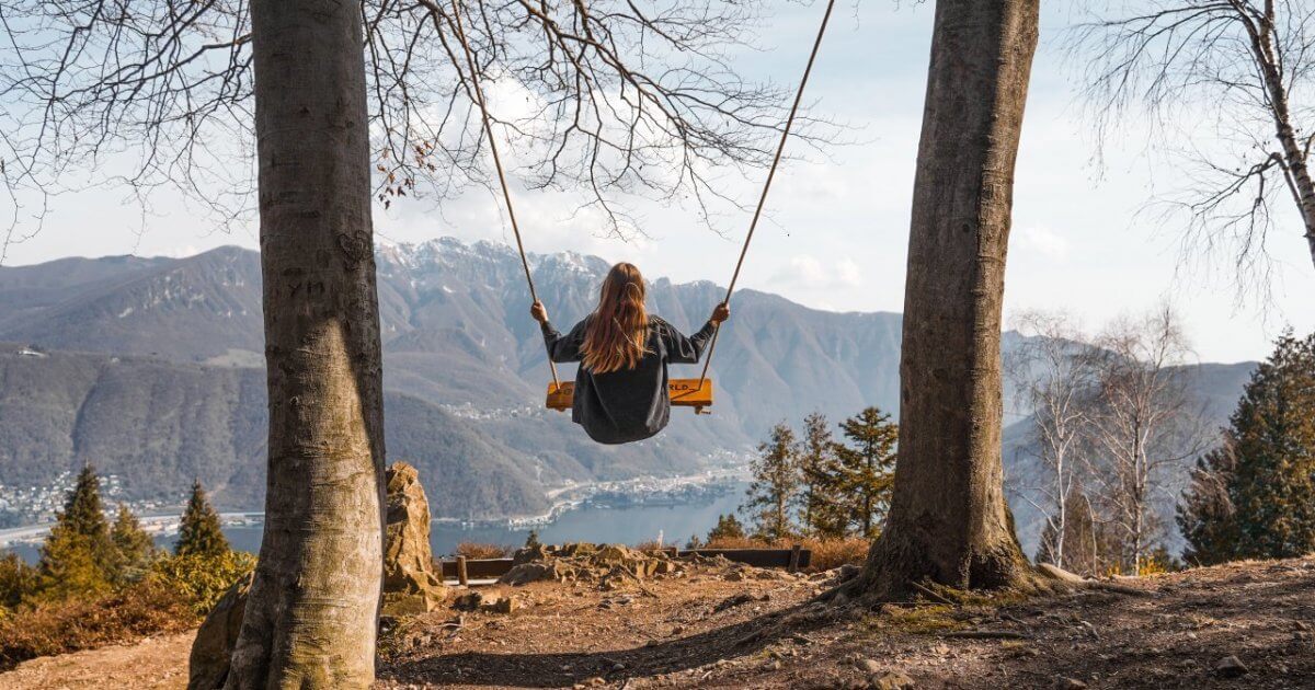 A girl swinging between two large trees with a stunning view of a lake and mountain range at Parco San Grato in the background.