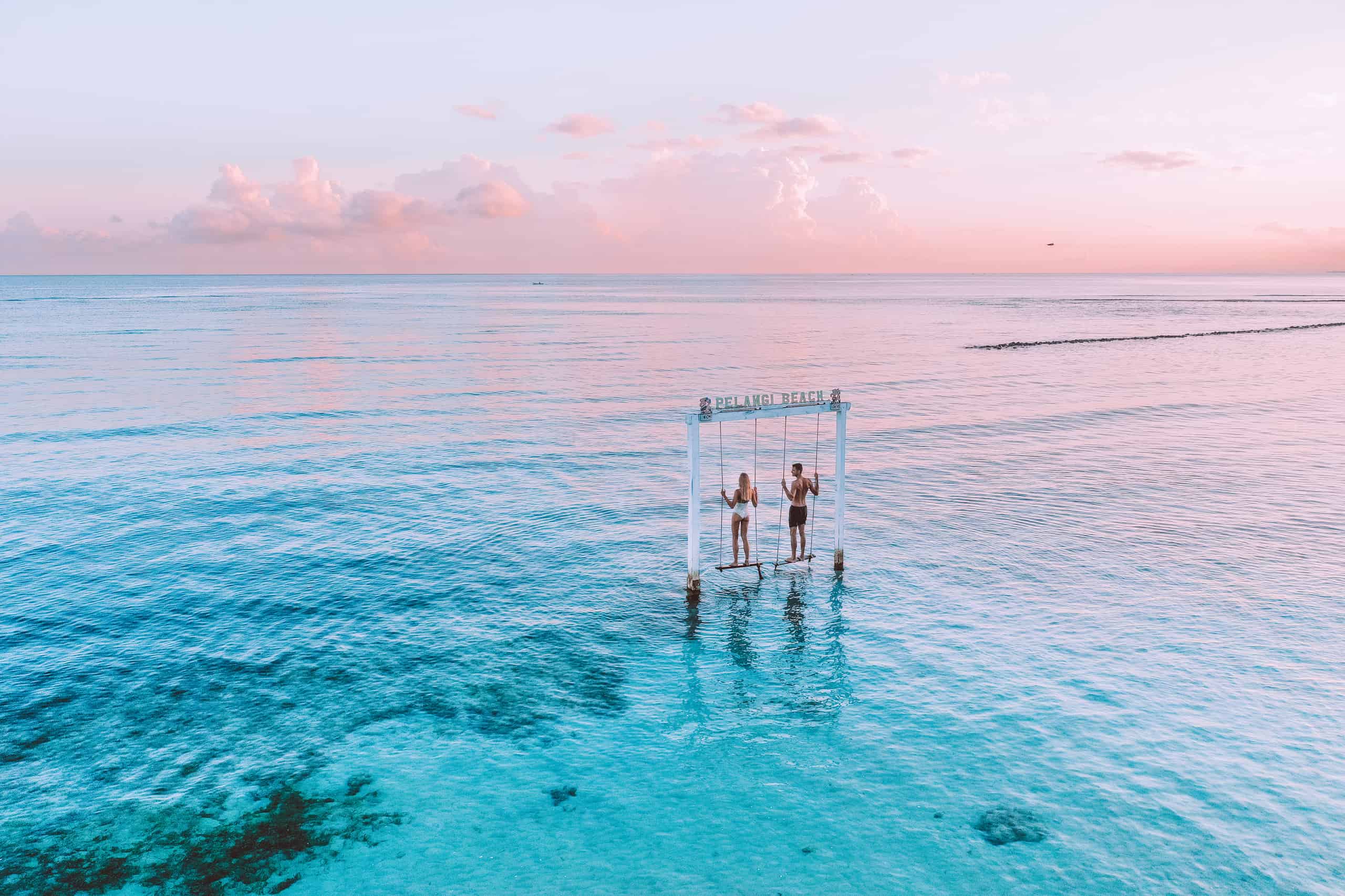 Two people stand on a white swing set labeled "Pelangi Beach" in crystal-clear turquoise waters against a pastel sunset sky.