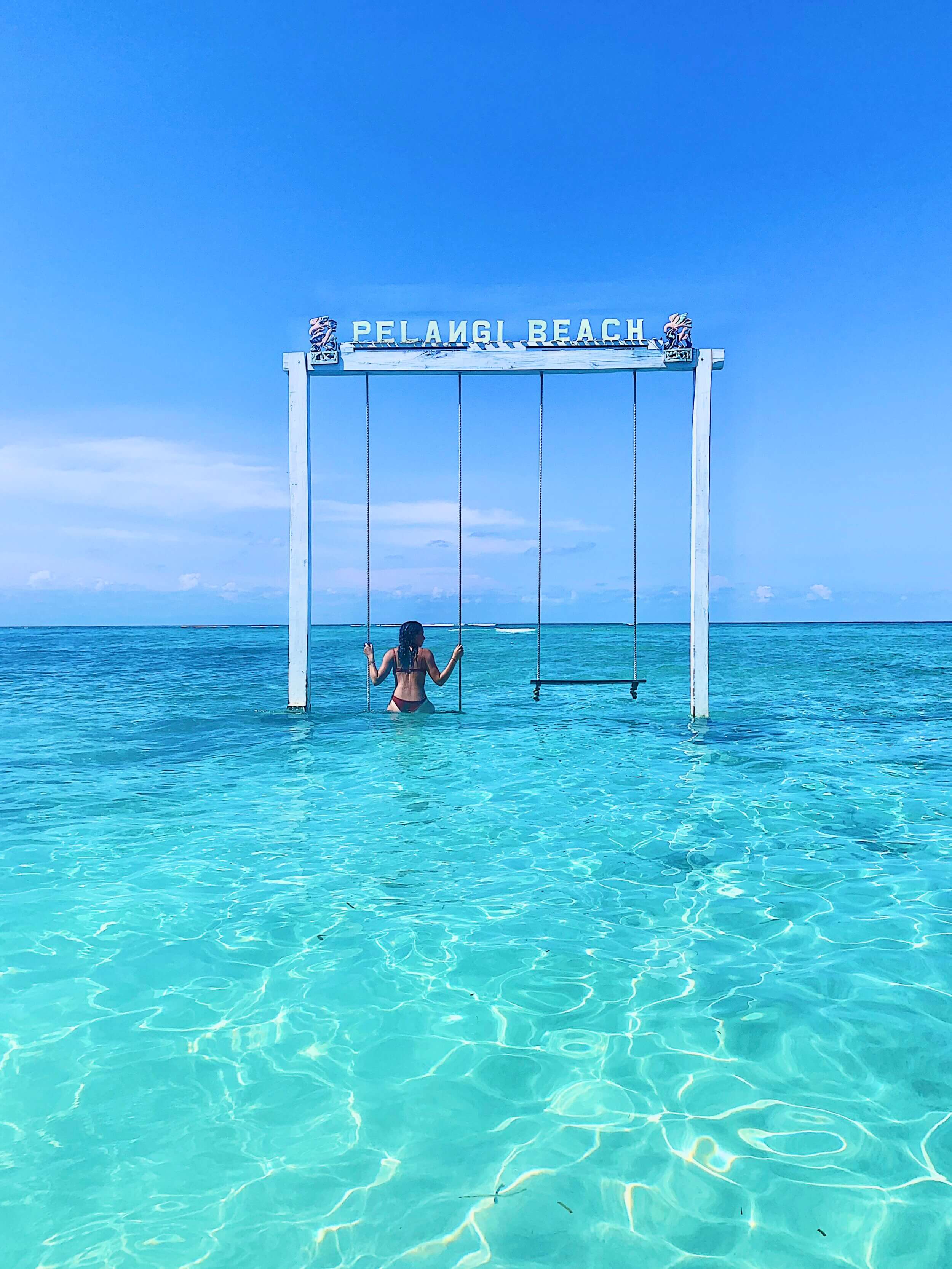 A woman in a bikini sits on a swing attached to a white frame labeled "Pelangi Beach" in shallow, crystal-clear turquoise waters on a sunny day.