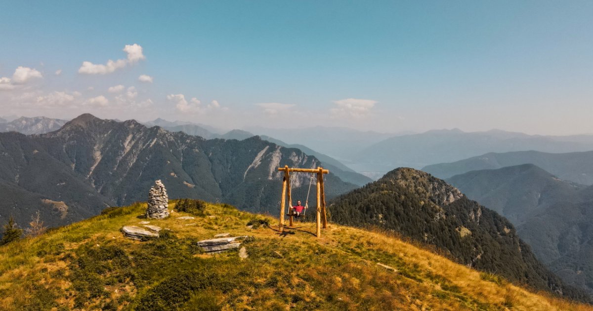 A wooden swing on a mountaintop overlooks a vast landscape of rugged peaks and valleys under a clear blue sky.