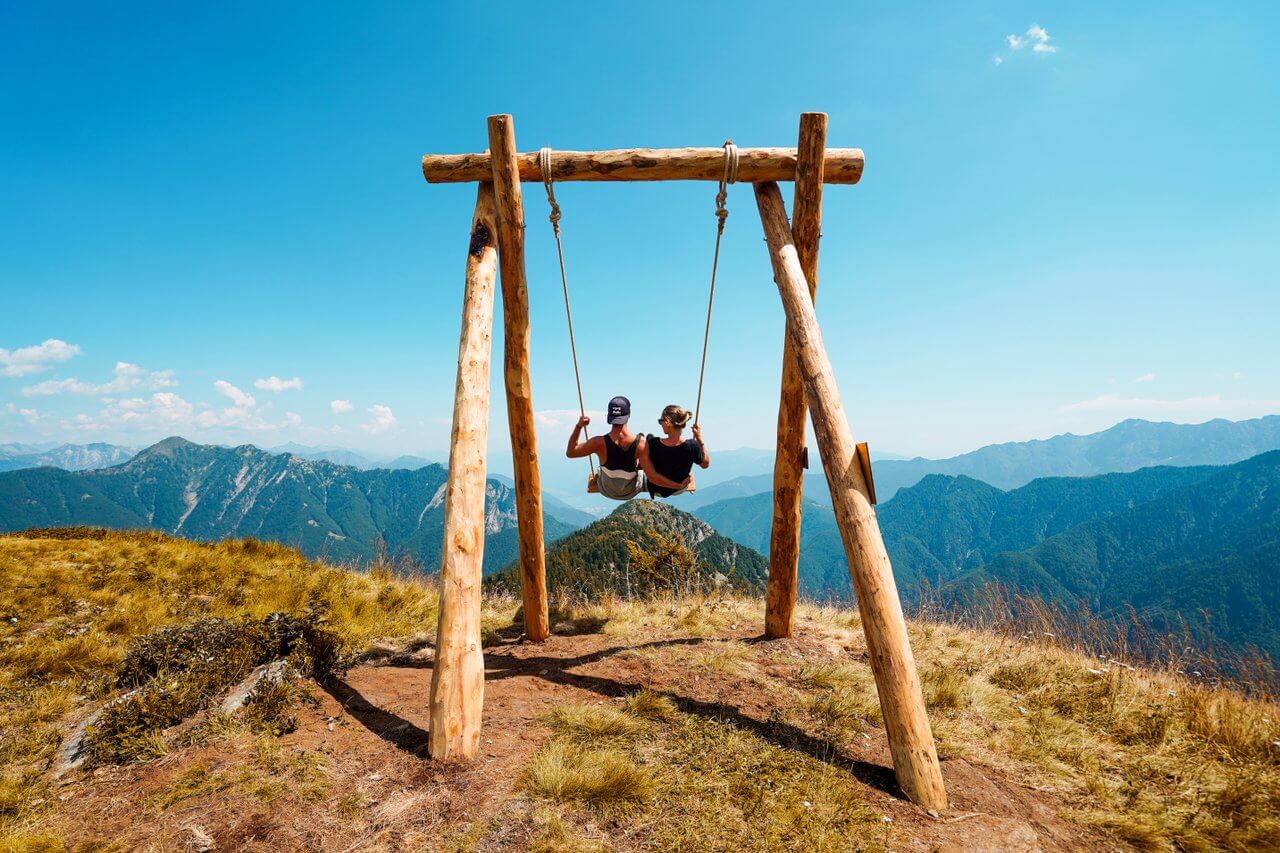 Two people sit on a rustic wooden swing perched atop a grassy mountain summit, enjoying panoramic views of distant mountain ranges.