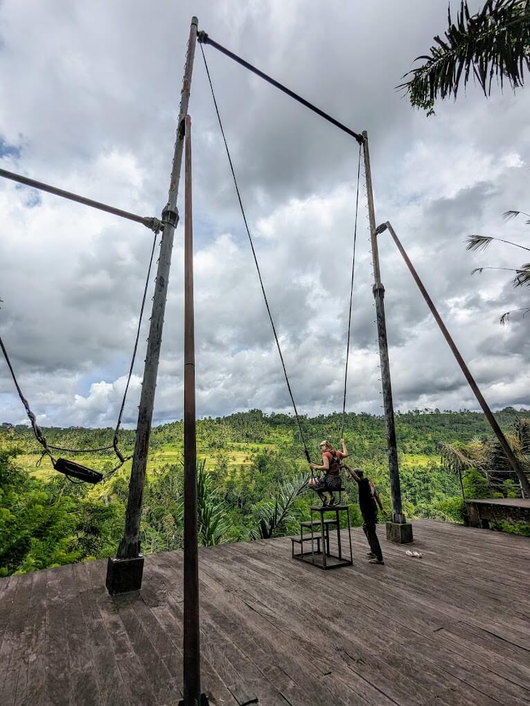 A person is seated on a large swing overlooking a lush green valley while being assisted by a guide, set against a dramatic cloudy sky.