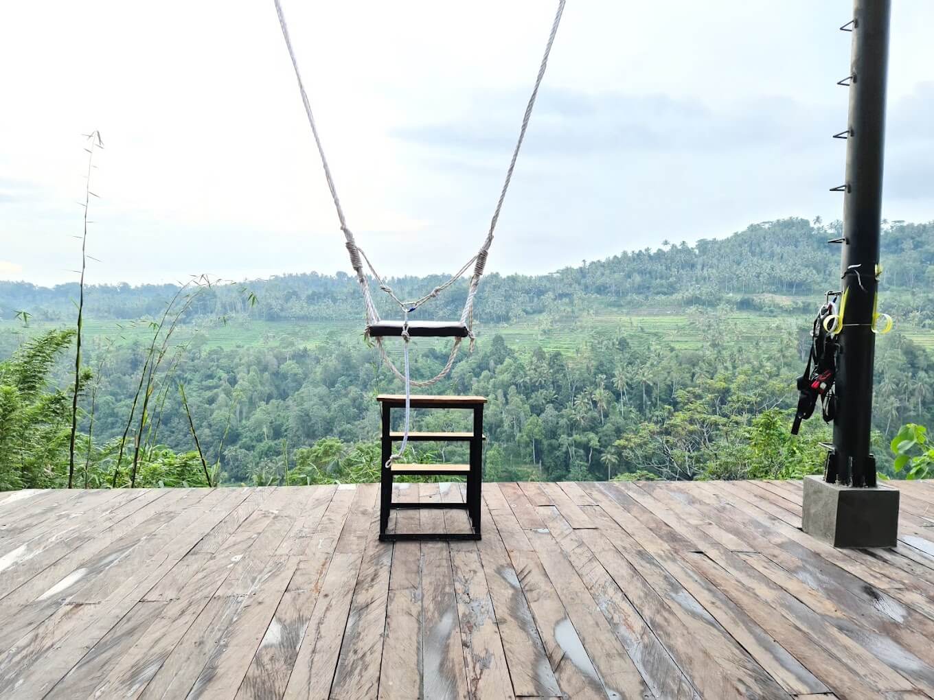 A wooden swing, framed by ropes, sits on a wooden platform facing a green valley with rolling hills and trees.