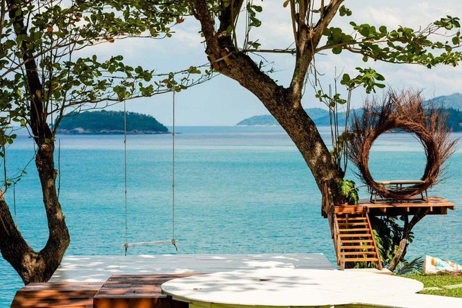 Ocean view with trees, wooden path to platform with swing. Rocky beach below. Cushioned daybeds for seating on grassy area.