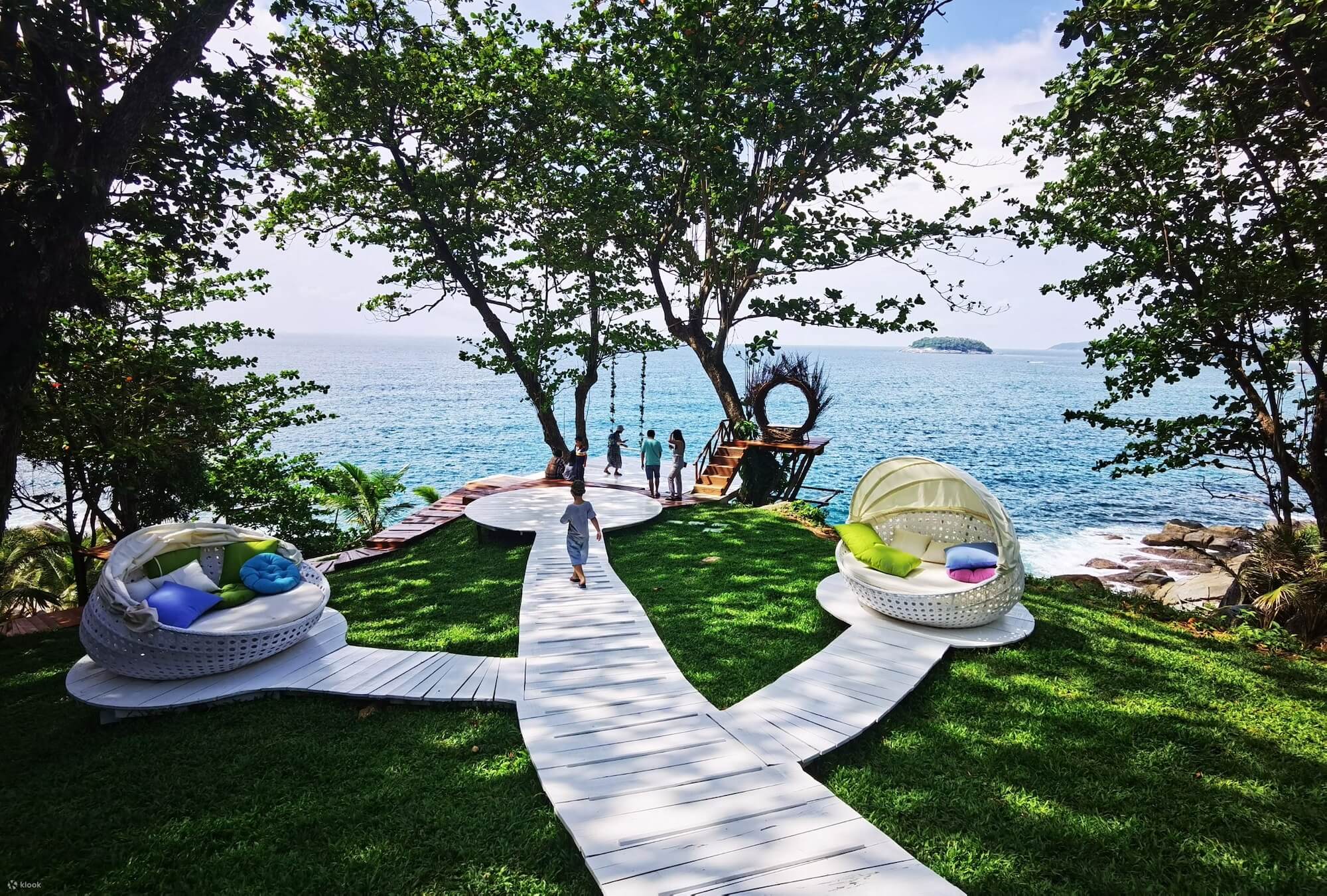 Ocean view with trees, wooden path to platform with swing. Rocky beach below. Cushioned daybeds for seating on grassy area.