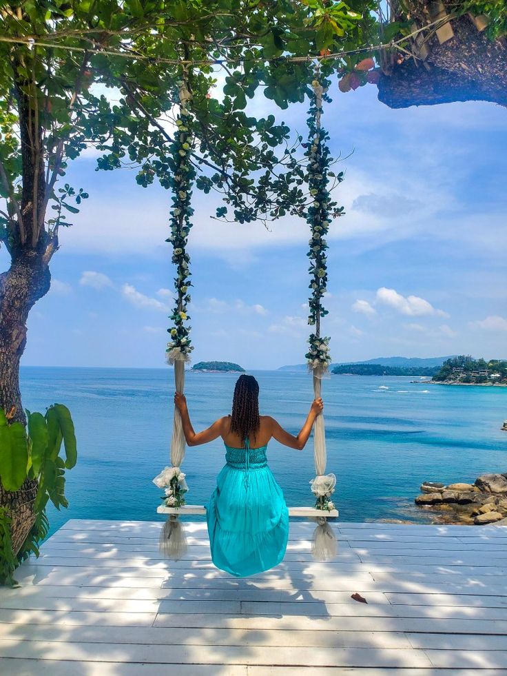 Woman in a turquoise dress sitting on a decorated swing at Rock Beach, facing a serene ocean view with small islands in the distance, surrounded by trees and greenery.