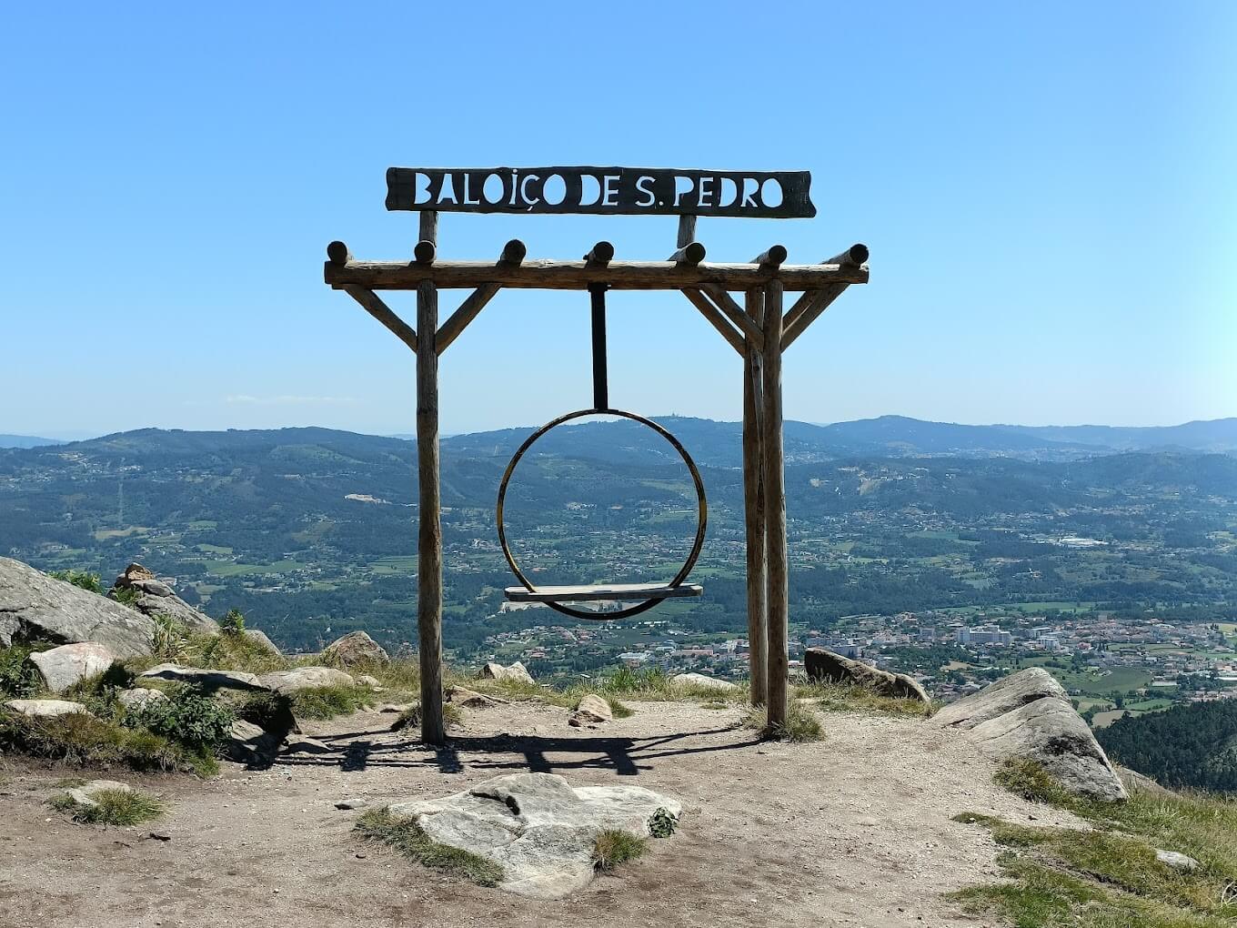 This image captures the São Pedro Fins swing with a panoramic view of green hills and a small town in the distance under a bright blue sky.