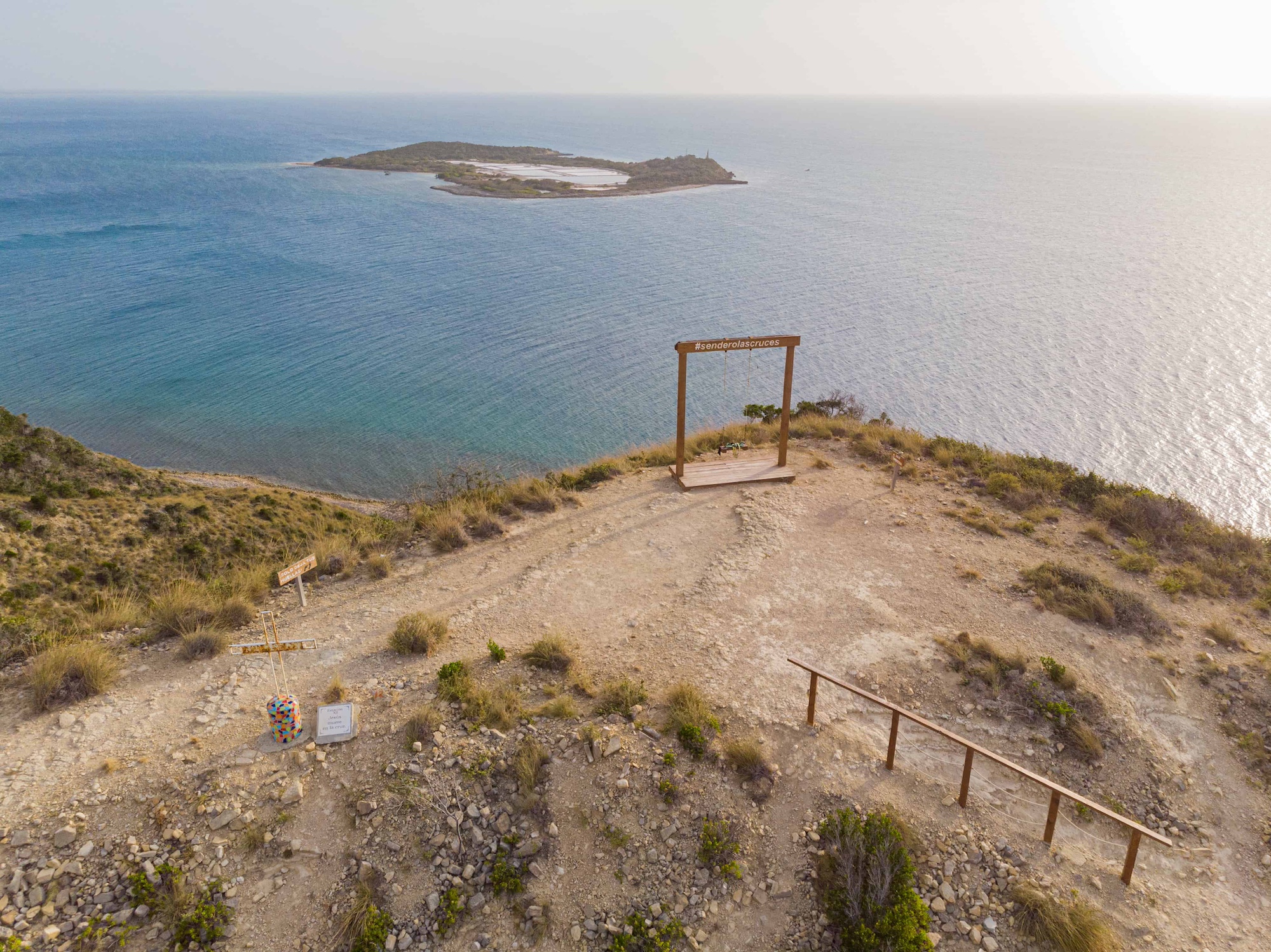 An aerial view of a wooden swing frame labeled "#senderolascruces" perched on a cliff overlooking the sea, with an island visible in the distance.