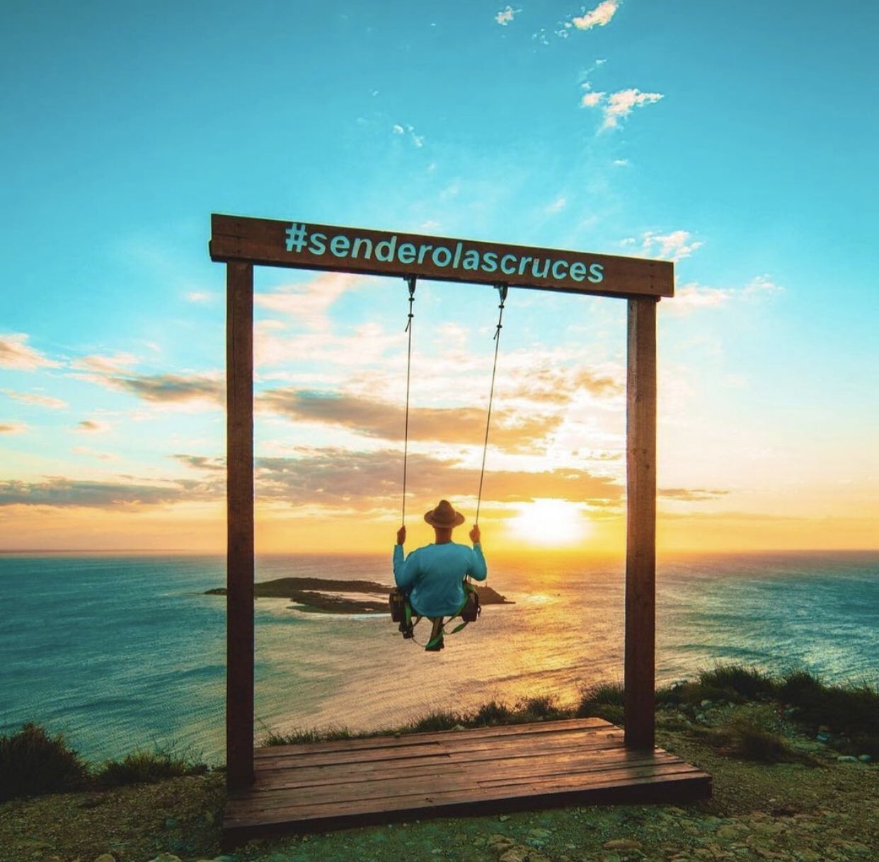 A person in a blue shirt and hat swings on a wooden frame labeled "#senderolascruces" against a stunning sunset backdrop overlooking the ocean and an island.