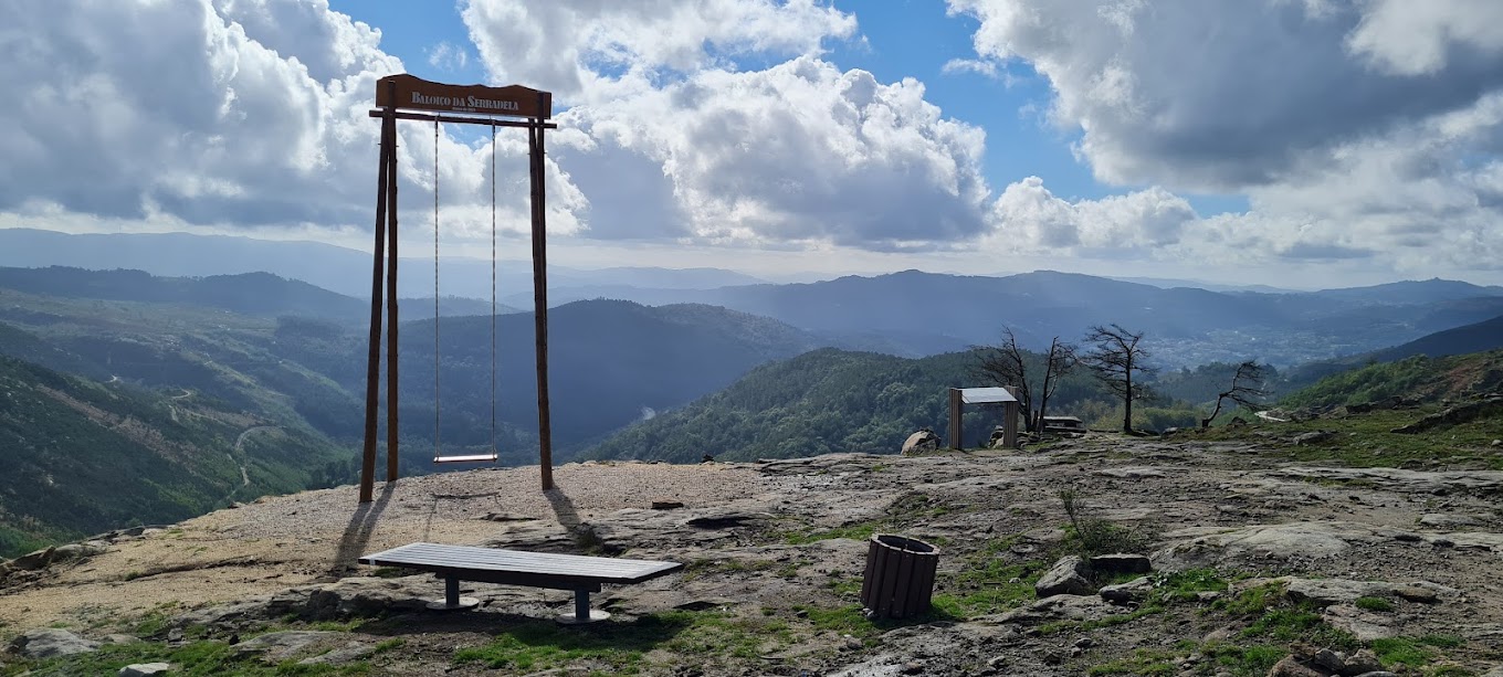 A tall wooden swing frame labeled "Baloico da Serradela" stands on a rocky mountaintop overlooking a vast, misty valley landscape, with a bench and trash bin nearby.