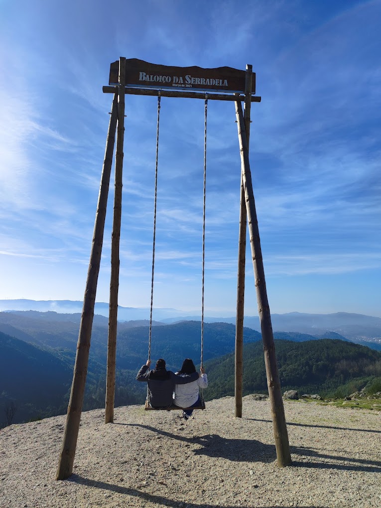 Two people sit on a large wooden swing labeled "Baloico da Serradela", overlooking a panoramic view of mountainous terrain under a blue sky.