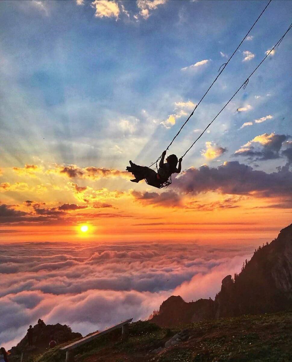 A woman swings on the Swing Above The Clouds, Turkey at sunset, with vibrant colors lighting the sky.