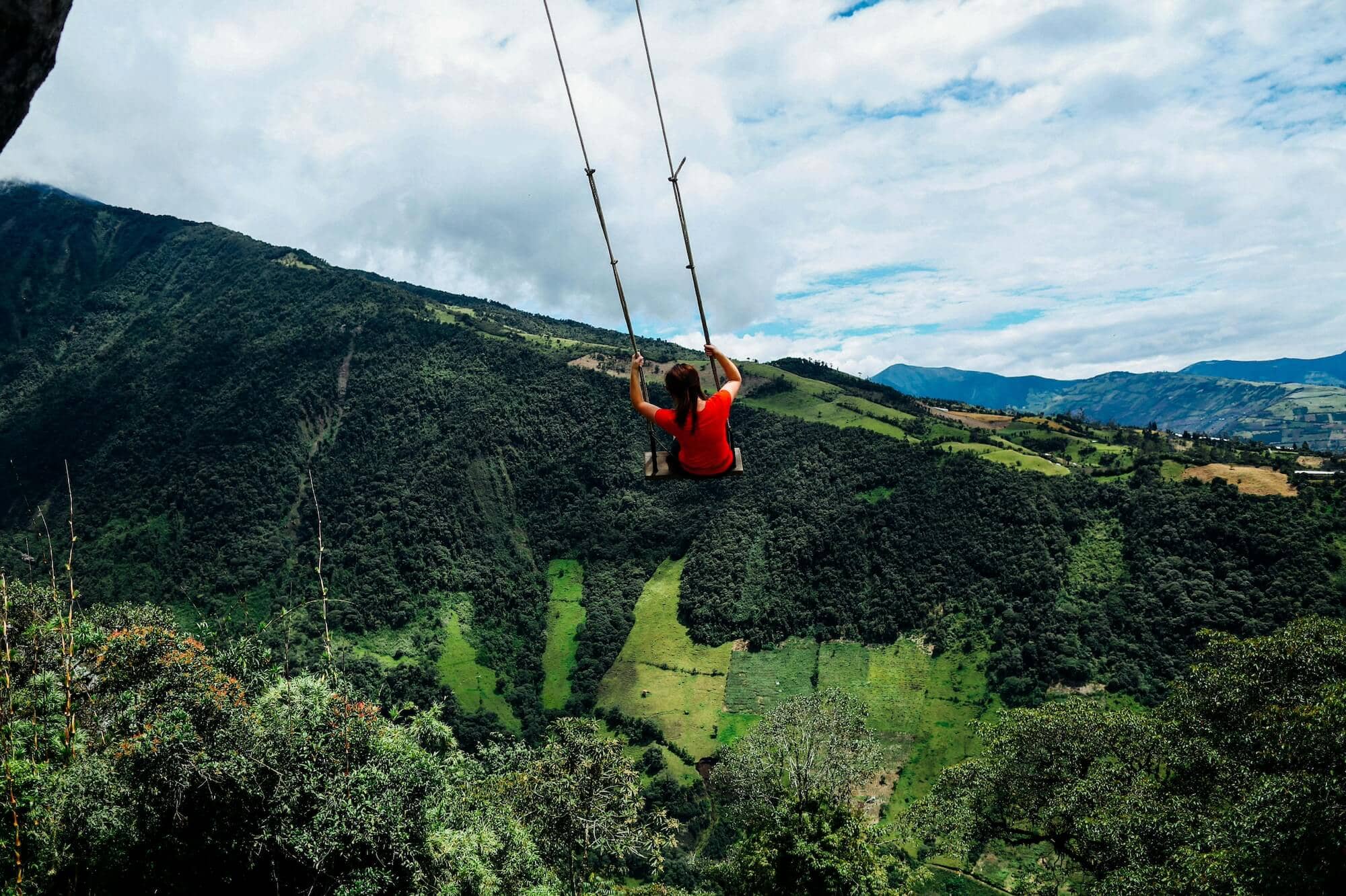 A woman in a red shirt is swinging on the Swing at the End of the World at the edge of a cliff, overlooking a vast valley with lush green forests and fields.