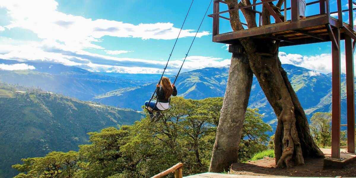 A woman is swinging on a Swing at the End of the World at the edge of a cliff, with a breathtaking view of mountains and lush greenery below.