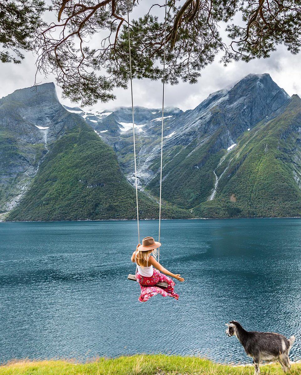 A person in a sun hat and pink skirt swings over a serene fjord, framed by towering mountains and pine branches, with a goat grazing in the foreground.