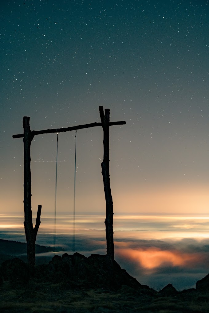 A rustic wooden swing frame silhouetted against a starry night sky with a colorful sunset on the horizon.