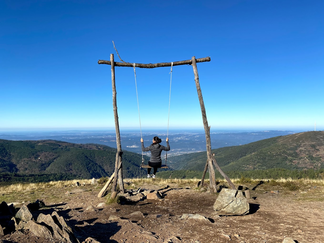 A person swings on a large wooden swing overlooking a vast mountainous landscape on a clear, sunny day.