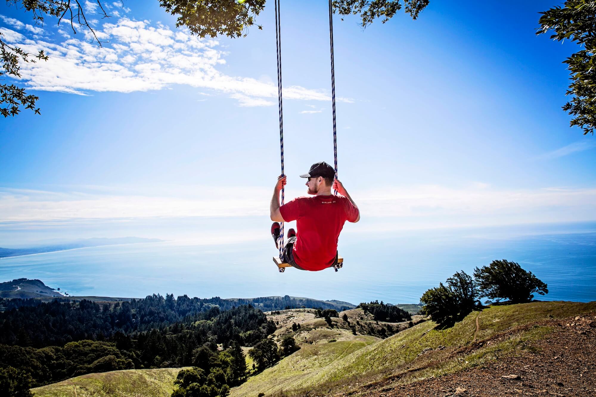 A man in a red shirt swings on the Trojan Point Swing, overlooking a coastal landscape with hills and ocean views on a sunny day.