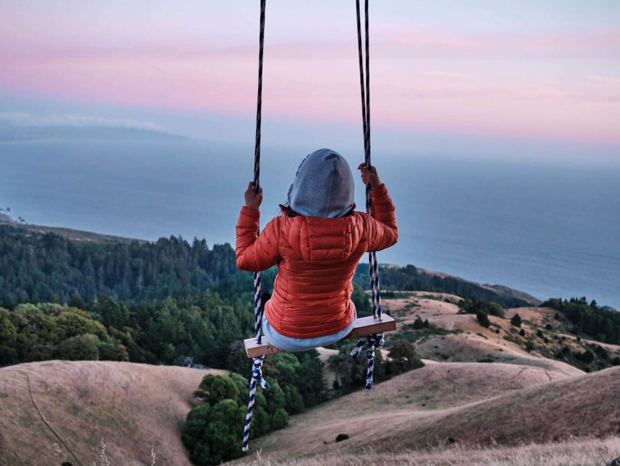 A woman in a red jacket swings on the Trojan Point Swing, overlooking hills, forest, and coastline at sunrise or sunset.