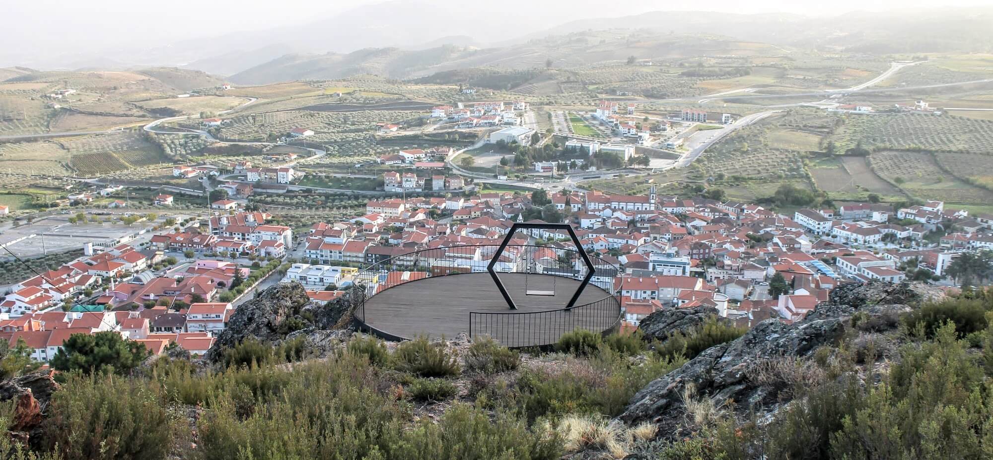 A scenic overlook above a town surrounded by rolling hills, olive groves, and farmland, with a hexagonal observation deck in the foreground.