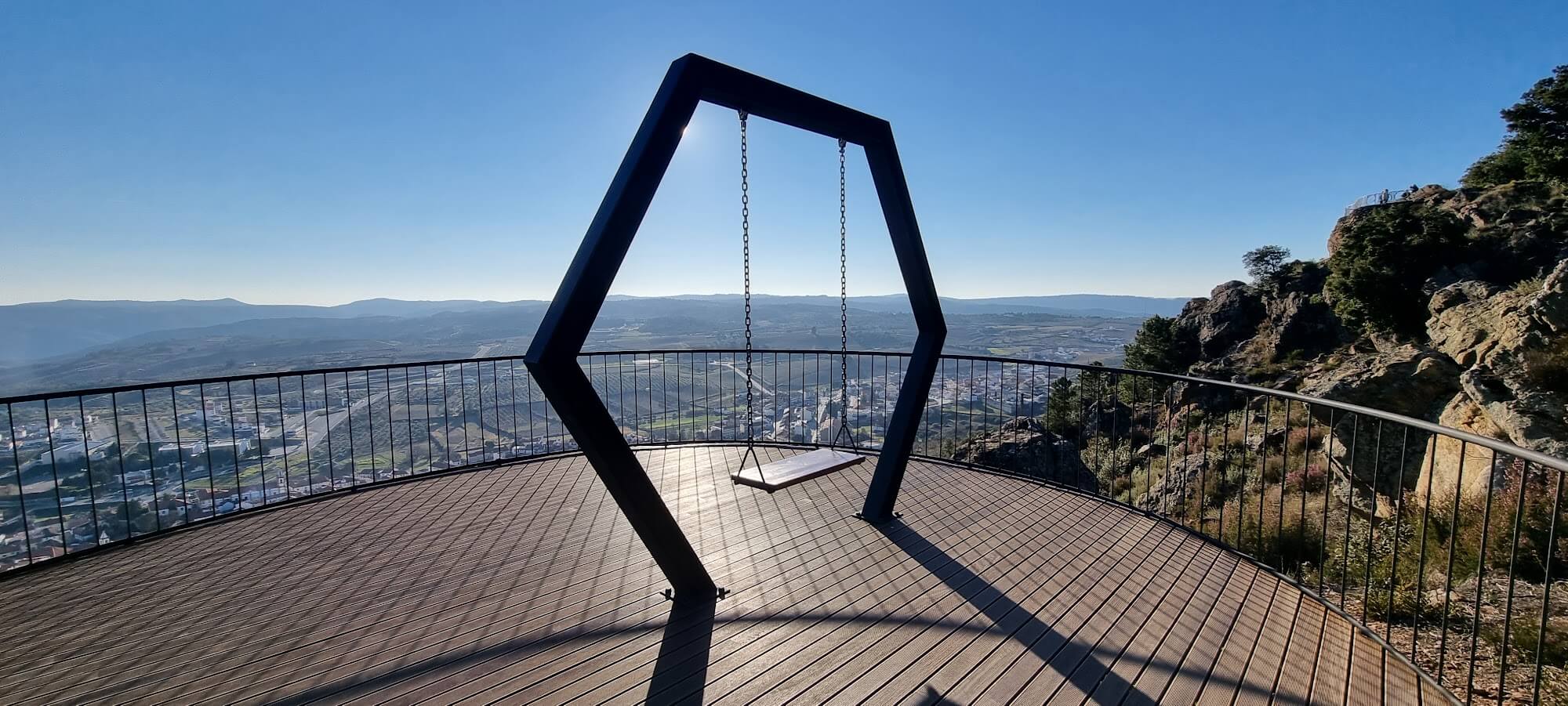 A swing hanging from a hexagonal frame on a wooden observation deck, offering a panoramic view of a town and distant mountains under a clear sky.